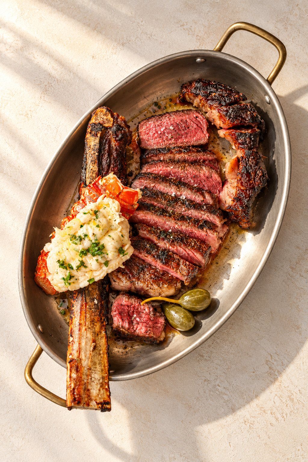 Sliced bone-in steak with a lobster tail and caperberries in a brass-handled pan